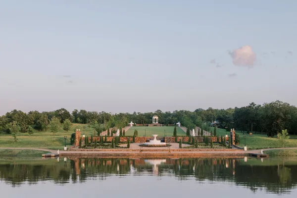 Wide landscape view of a European-style estate featuring manicured gardens, a central pavilion, and a reflecting pond.