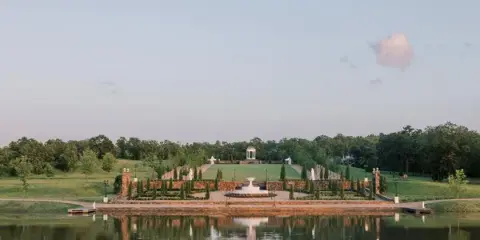 Wide landscape view of a European-style estate featuring manicured gardens, a central pavilion, and a reflecting pond.