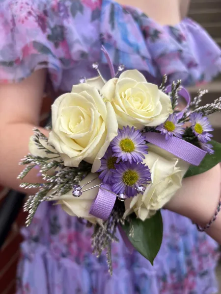 A hand holding a formal dance bouquet featuring white roses, purple daisies, and a purple ribbon.