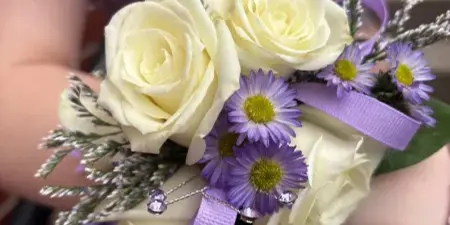 A hand holding a formal dance bouquet featuring white roses, purple daisies, and a purple ribbon.