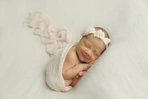 A sleeping newborn wrapped in a cream knit wrap with a floral headband, photographed on a soft white backdrop.