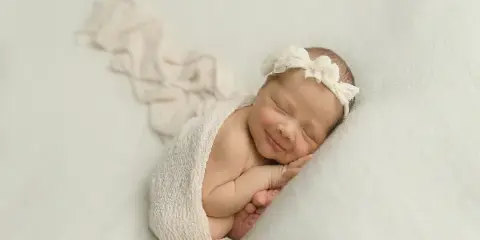 A sleeping newborn wrapped in a cream knit wrap with a floral headband, photographed on a soft white backdrop.