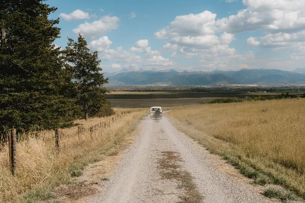 A gravel farm road runs through open grassland with a vehicle approaching, bordered by trees and a fence, with distant mountains under a partly cloudy sky.