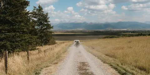 A gravel farm road runs through open grassland with a vehicle approaching, bordered by trees and a fence, with distant mountains under a partly cloudy sky.