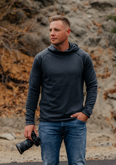 A man in a dark hoodie and jeans stands outdoors holding a professional camera.