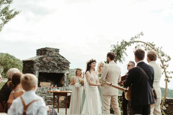 A bride and groom exchanging vows during an outdoor wedding ceremony on a stone terrace with a fireplace and mountain views.