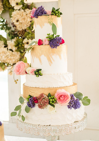 A three-tiered white wedding cake decorated with pink and purple flowers and greenery, displayed on a silver stand.