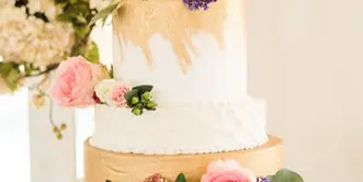 A three-tiered white wedding cake decorated with pink and purple flowers and greenery, displayed on a silver stand.