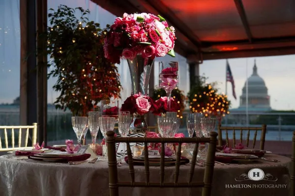A formal event table setting with bamboo chairs, white linens, and floral centerpieces, set against a backdrop of the U.S. Capitol building.