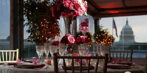 A formal event table setting with bamboo chairs, white linens, and floral centerpieces, set against a backdrop of the U.S. Capitol building.