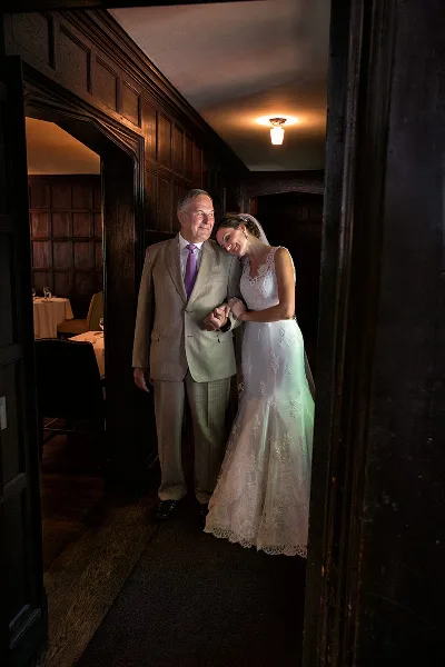 A bride in a lace gown leaning affectionately against her father in a suit, standing in a wood-paneled hallway.