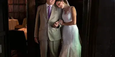 A bride in a lace gown leaning affectionately against her father in a suit, standing in a wood-paneled hallway.