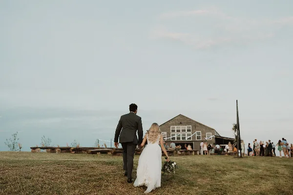 A bride and groom walking across a grassy hilltop toward a rustic barn wedding venue with guests gathered nearby.
