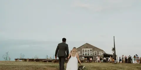 A bride and groom walking across a grassy hilltop toward a rustic barn wedding venue with guests gathered nearby.