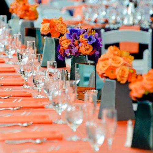 A long banquet table set with orange napkins, glassware, and floral centerpieces.