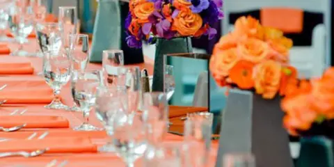 A long banquet table set with orange napkins, glassware, and floral centerpieces.
