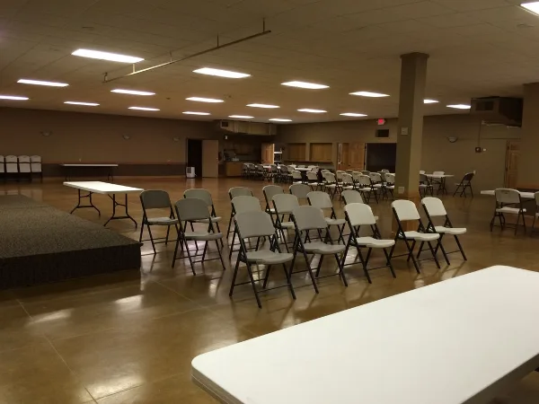 Interior of a large event hall with rows of folding chairs, tables, and a stage area.