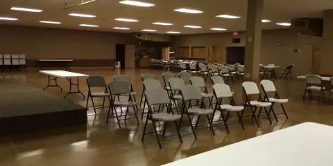 Interior of a large event hall with rows of folding chairs, tables, and a stage area.