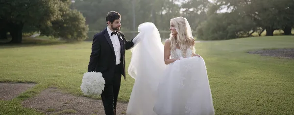 A bride and groom standing in a grassy field, with the groom lifting the bride's veil in a candid, documentary-style shot.