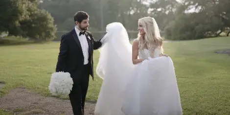 A bride and groom standing in a grassy field, with the groom lifting the bride's veil in a candid, documentary-style shot.
