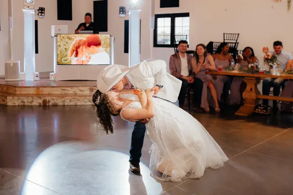 A DJ at a wedding reception booth with a laptop and screen, while a couple performs a dip on the dance floor in front of seated guests.