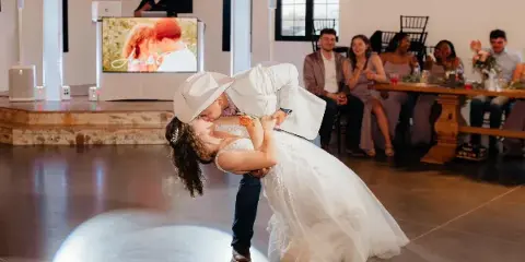 A DJ at a wedding reception booth with a laptop and screen, while a couple performs a dip on the dance floor in front of seated guests.