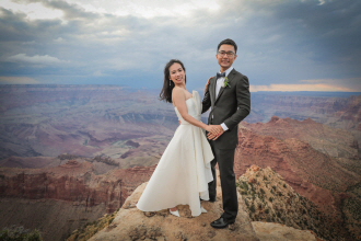 A bride and groom in formal wedding attire standing on a rocky ledge overlooking the Grand Canyon.