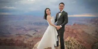 A bride and groom in formal wedding attire standing on a rocky ledge overlooking the Grand Canyon.