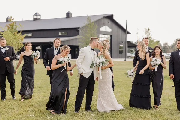 A wedding party posing on a lawn in front of a large, modern black barn wedding venue.