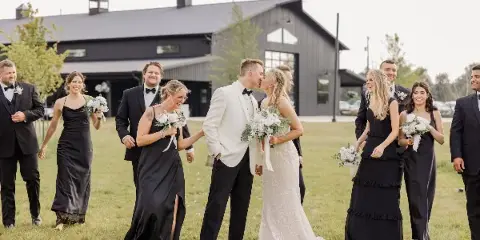 A wedding party posing on a lawn in front of a large, modern black barn wedding venue.