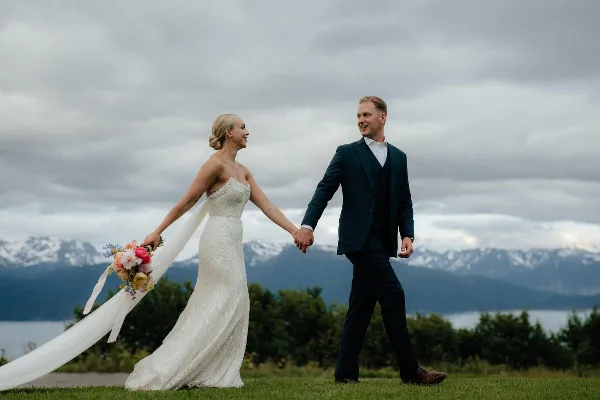 A bride and groom walking hand-in-hand on a grassy field with a mountain and lake backdrop.