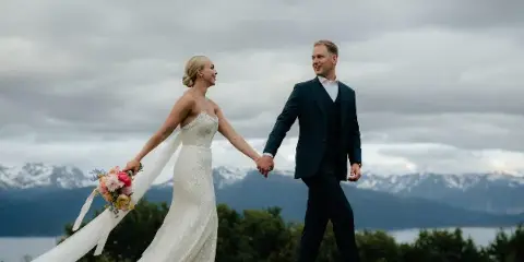 A bride and groom walking hand-in-hand on a grassy field with a mountain and lake backdrop.