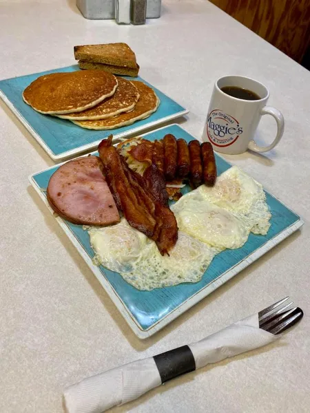 Breakfast platter with eggs, bacon, sausage, ham, and pancakes on a blue plate next to a coffee mug with the Maggie's Cafe & Catering logo.