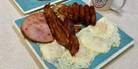 Breakfast platter with eggs, bacon, sausage, ham, and pancakes on a blue plate next to a coffee mug with the Maggie's Cafe & Catering logo.