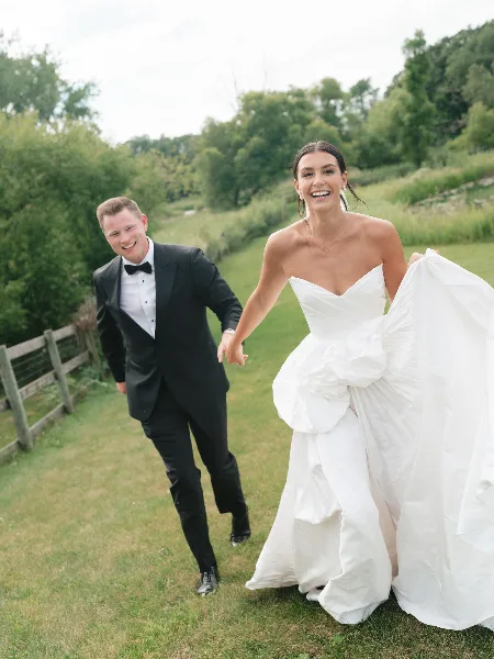 Bride in a white gown and groom in a black tuxedo walking hand-in-hand on a grassy field.