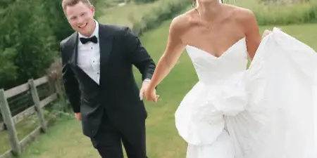 Bride in a white gown and groom in a black tuxedo walking hand-in-hand on a grassy field.