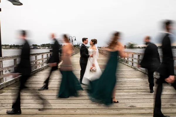 A bride and groom standing on a wooden pier with motion-blurred wedding party members in the foreground.