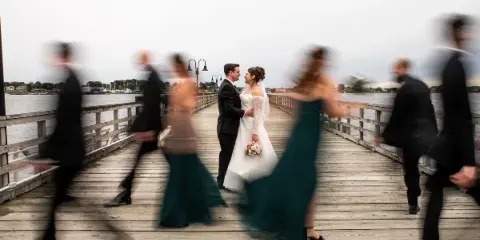 A bride and groom standing on a wooden pier with motion-blurred wedding party members in the foreground.