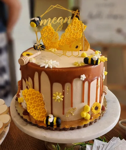 A custom decorated cake featuring honeycomb panels, fondant bees, and a honey dipper on a marble cake stand.
