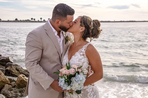 A bride and groom kissing on a beach, with the bride holding a bouquet of pink roses and blue hydrangeas.