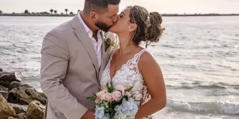 A bride and groom kissing on a beach, with the bride holding a bouquet of pink roses and blue hydrangeas.