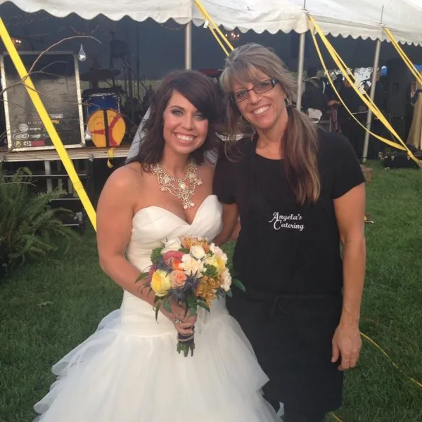 A bride in a white wedding dress holding a bouquet poses next to a smiling catering staff member wearing a black apron with 'Angela's Catering' written on it.