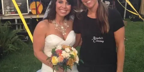 A bride in a white wedding dress holding a bouquet poses next to a smiling catering staff member wearing a black apron with 'Angela's Catering' written on it.
