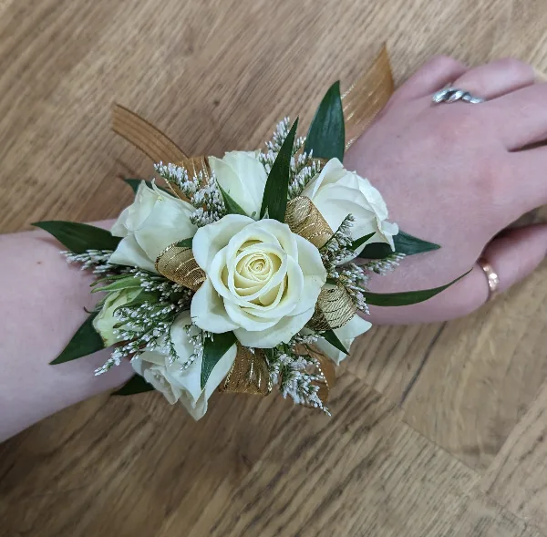 A white rose and greenery floral corsage with gold ribbon accents worn on a wrist.