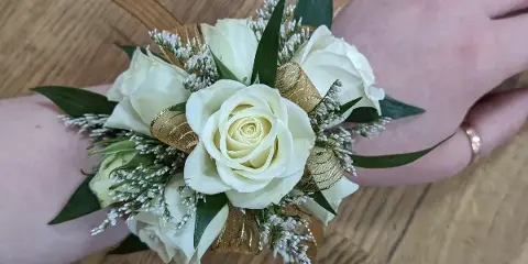 A white rose and greenery floral corsage with gold ribbon accents worn on a wrist.