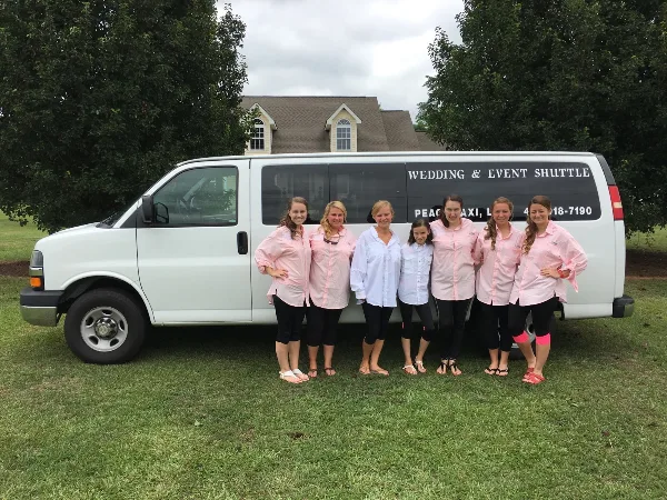 A group of women in matching pink shirts standing in front of a white Peach Taxi van labeled 'Wedding & Event Shuttle'.