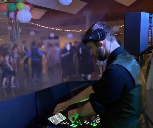 A DJ wearing headphones operates a mixing console at a wedding reception while guests dance in the background.