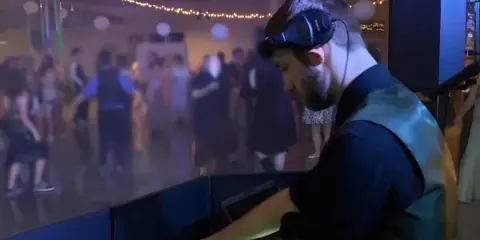 A DJ wearing headphones operates a mixing console at a wedding reception while guests dance in the background.