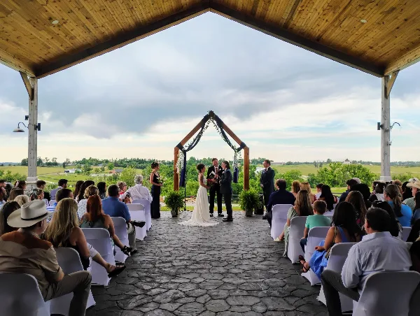 A wedding ceremony in progress under a large wooden awning with a stone floor, surrounded by seated guests and overlooking a rural landscape.