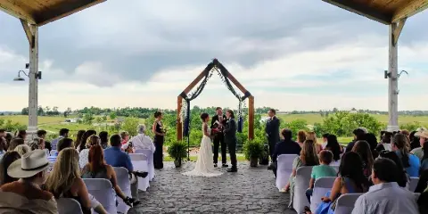 A wedding ceremony in progress under a large wooden awning with a stone floor, surrounded by seated guests and overlooking a rural landscape.
